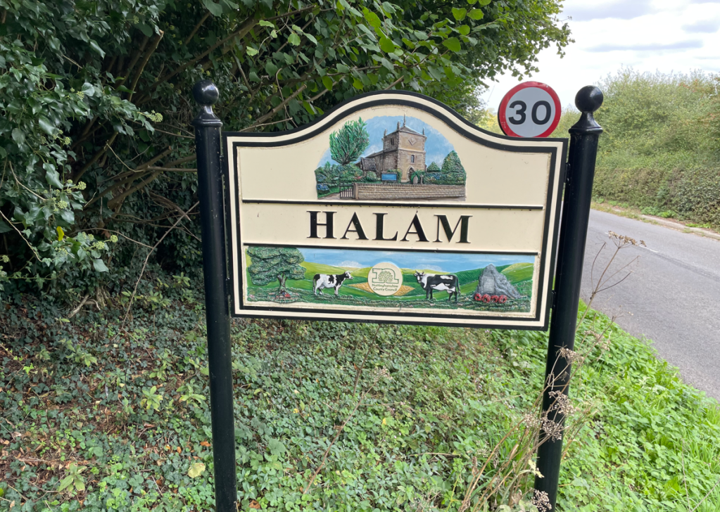 Sign for the village of Halam, featuring an illustration of a church, trees, and a cow, next to a speed limit sign.