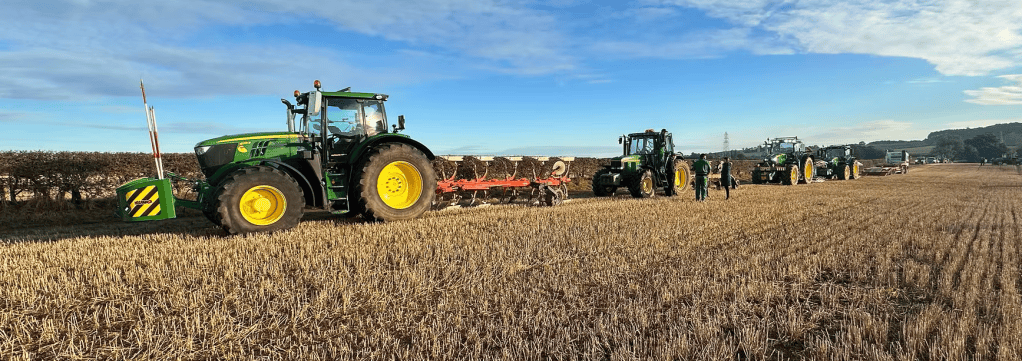 Three green tractors working in a field, with farm workers visible beside them and a clear blue sky above.