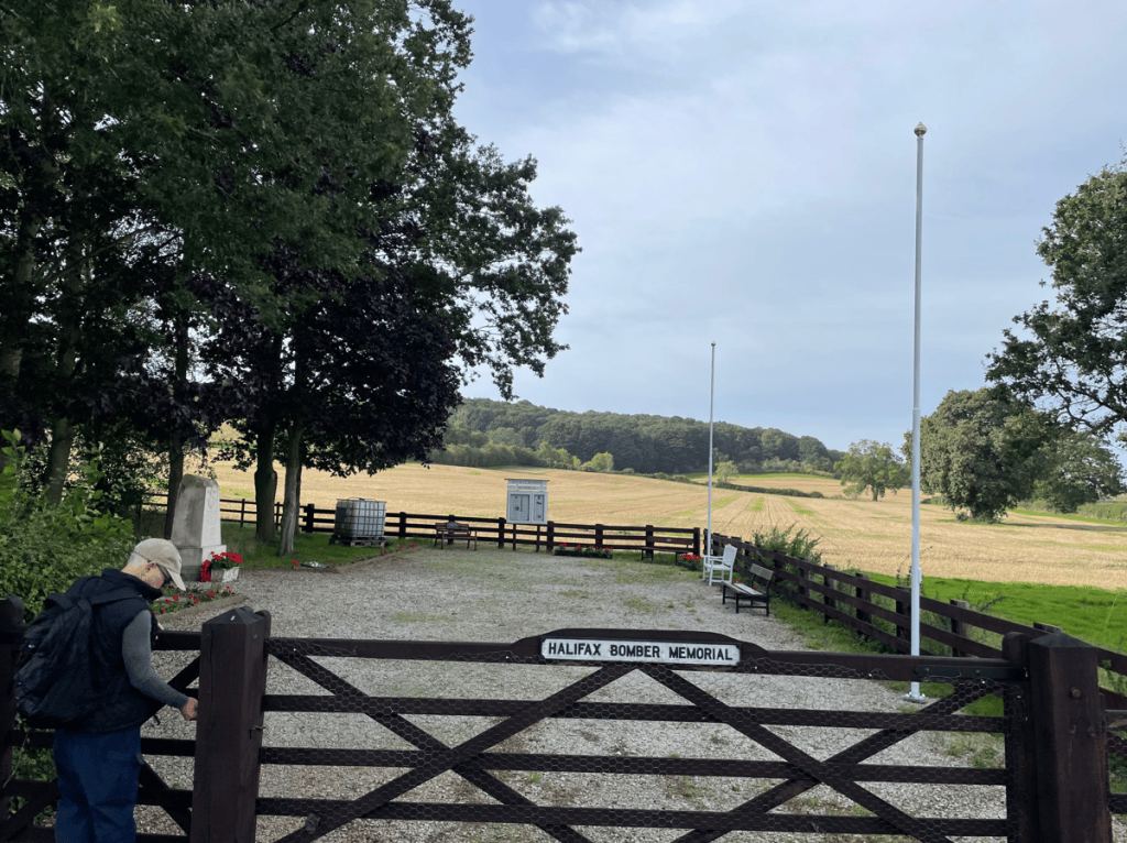 Entrance to the Halifax Bomber Memorial, featuring a wooden gate, flags, and a visitor near the entrance, surrounded by trees and open fields.