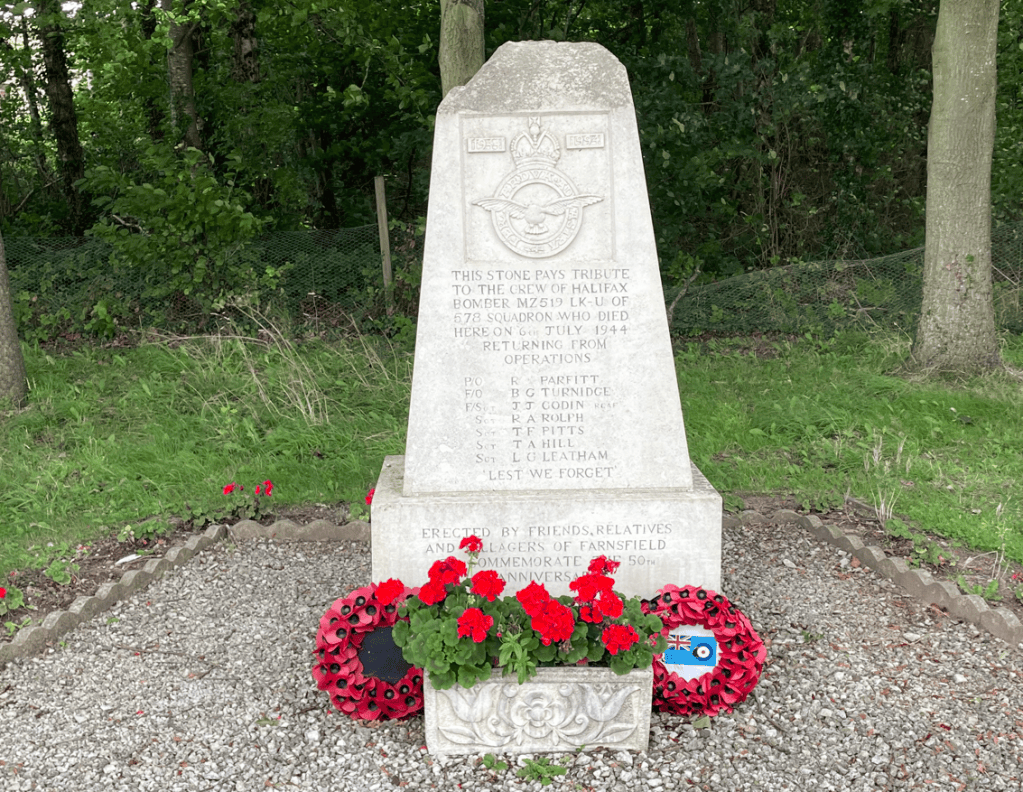A memorial stone dedicated to the crew of Halifax bomber MZ519 LK-UI, 578 Squadron, who died on 6th July 1944, surrounded by red flowers and remembrance wreaths.