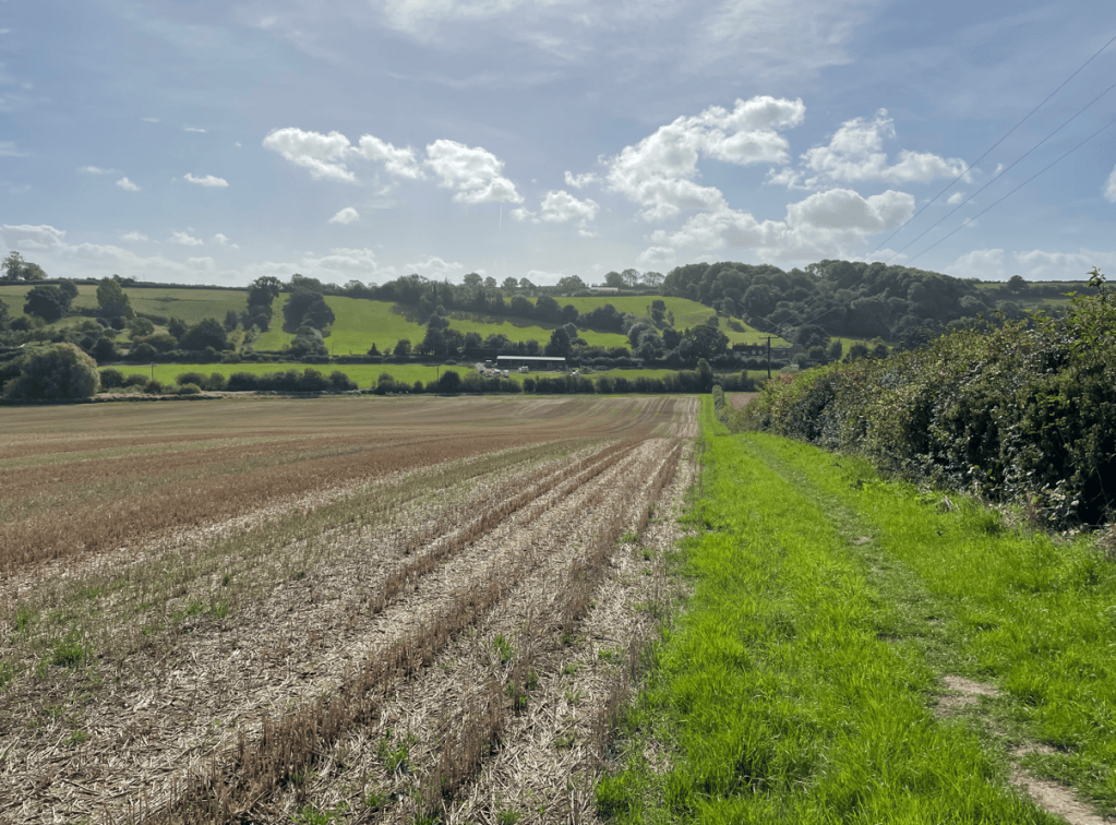 A picturesque view of a rural landscape showcasing a harvested field leading to lush greenery and rolling hills under a partly cloudy sky.