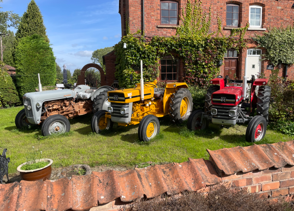 Three vintage tractors in a garden setting: a grey tractor, a yellow tractor, and a red tractor, with a brick house and greenery in the background.