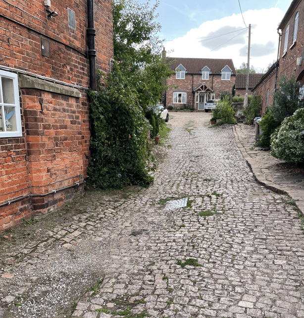 A narrow, cobbled alleyway lined with red-brick buildings and greenery, leading towards a house with a gabled roof and parked cars. The scene is set under a partly cloudy sky.