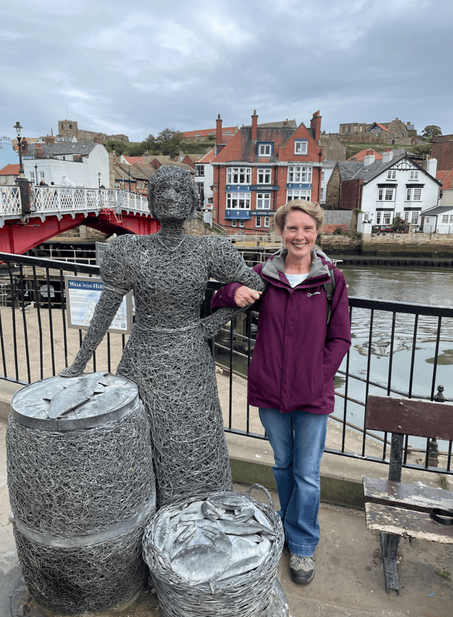 A woman standing next to a wire sculpture of a female figure, with barrels nearby, by a riverside in a quaint village.