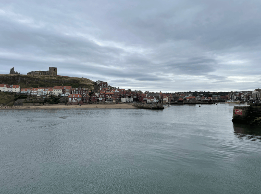 A view of a coastal town with red-roofed buildings lining the shore, under a cloudy sky. A hill with a historic church and castle ruins is visible in the background, while boats are seen in the water near the harbour.