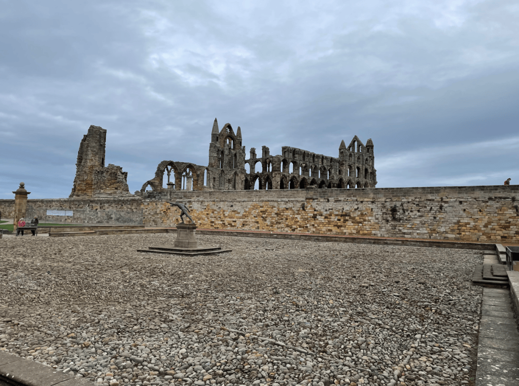 Ruins of a historic abbey against a cloudy sky, with a stone wall and a gravel area in the foreground.