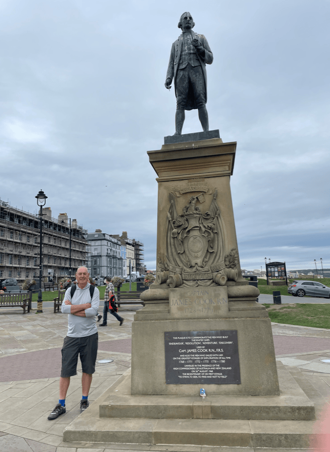 A man stands beside a statue of Captain James Cook, which is located on a plinth with a plaque, set against a cloudy sky and urban backdrop.