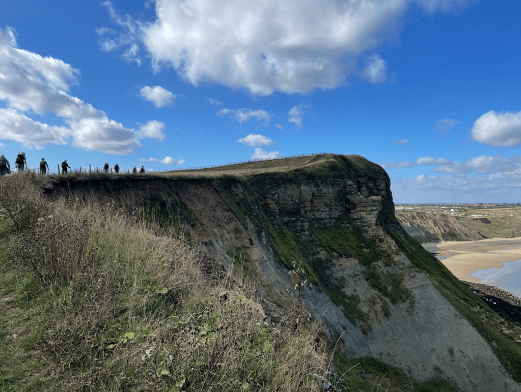 A scenic view of a coastal cliff with a walking path at the top, where several people are seen walking. The cliff features grassy vegetation and steep slopes, set against a blue sky with scattered clouds.
