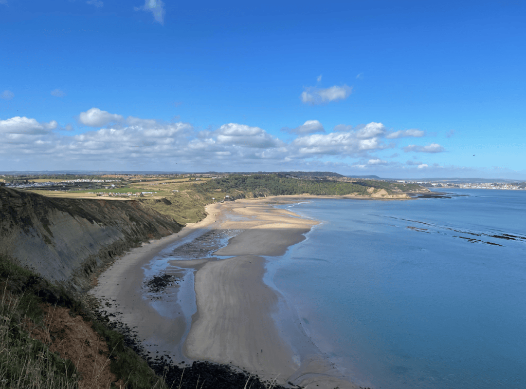 A scenic view of a sandy beach bordered by cliffs under a clear blue sky with scattered clouds.