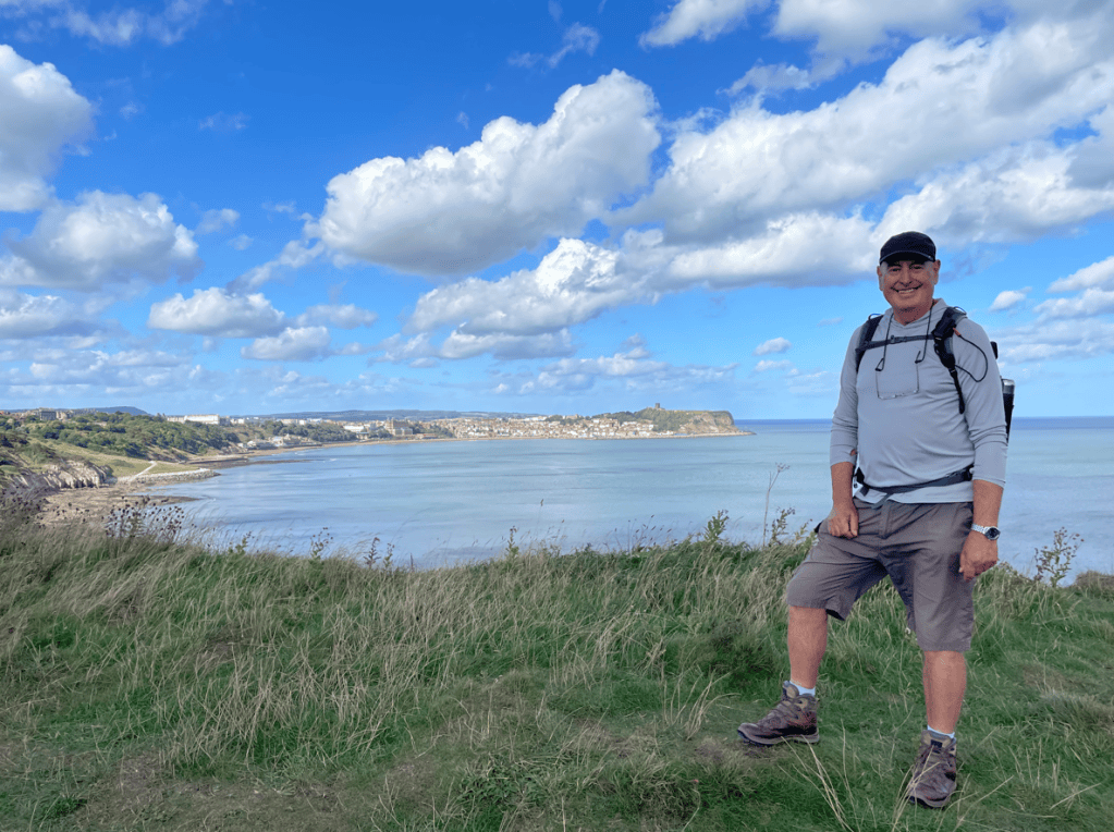 A man standing on a grassy hill overlooking the coastline with a blue sky and clouds in the background.