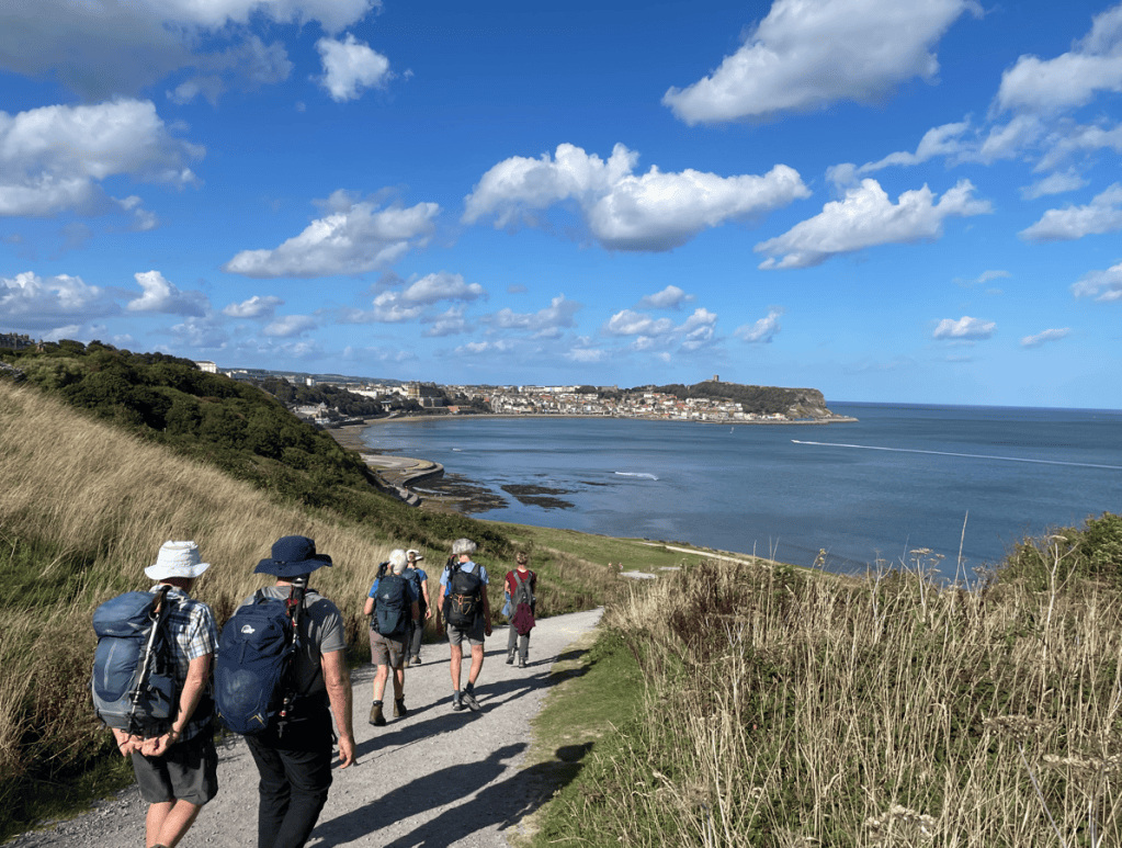 A group of hikers walking along a coastal path with views of the sea and a town in the background, under a blue sky with fluffy clouds.