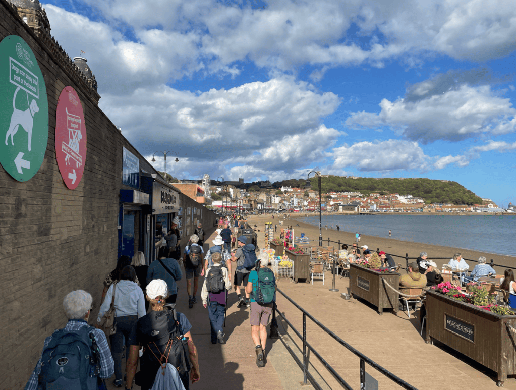 A busy seaside promenade with people walking along the pathway, shops on the left side, and benches with flowers facing the beach. A hill covered in greenery is visible in the background, under a partly cloudy sky.