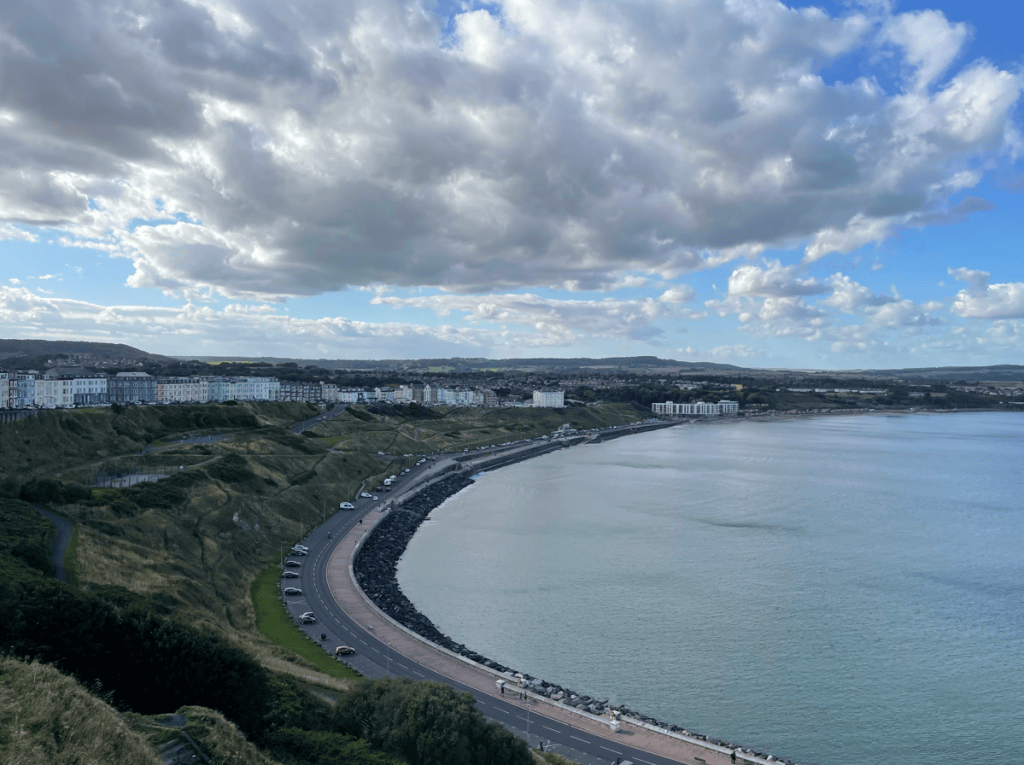 A scenic coastal view featuring a curving road alongside the sea, with green cliffs in the foreground and a town of white buildings in the background under a partly cloudy sky.