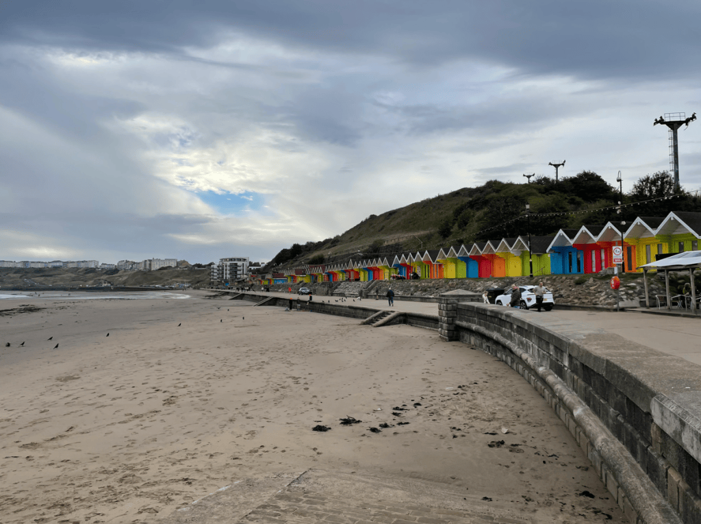 A beach scene featuring brightly coloured beach huts along the shoreline, with dark clouds overhead and a sandy beach in the foreground.