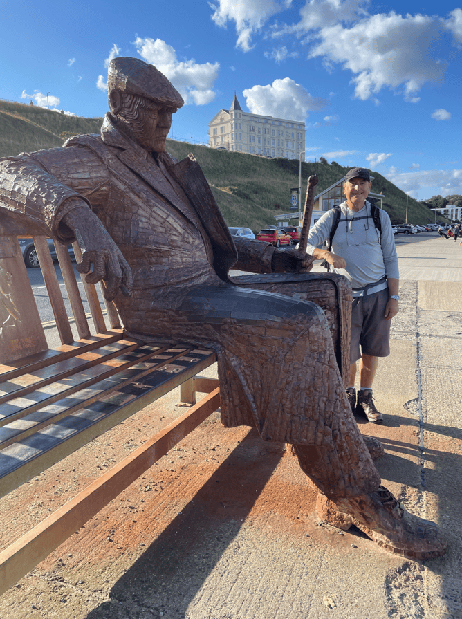 A man stands next to a large bronze statue of a seated figure on a bench, set against a backdrop of blue skies and clouds.