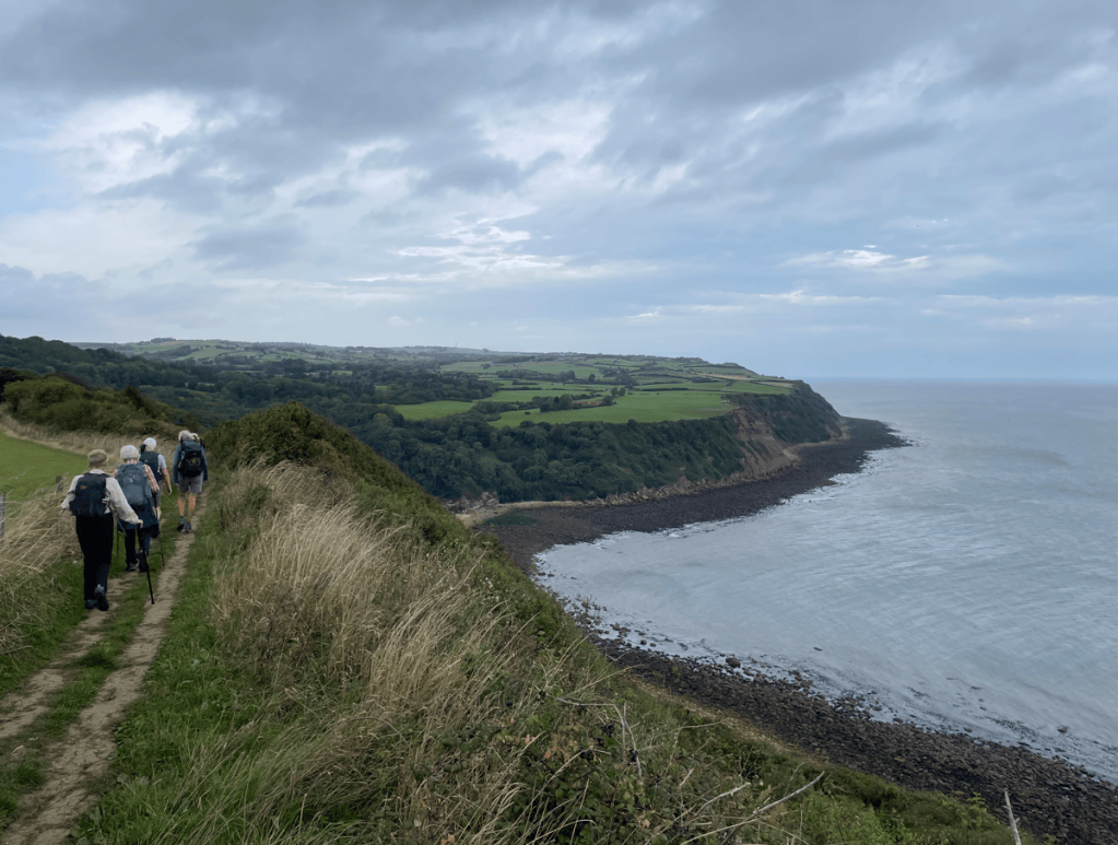 A group of hikers walking along a coastal path with cliffs and the sea visible in the background under a cloudy sky.