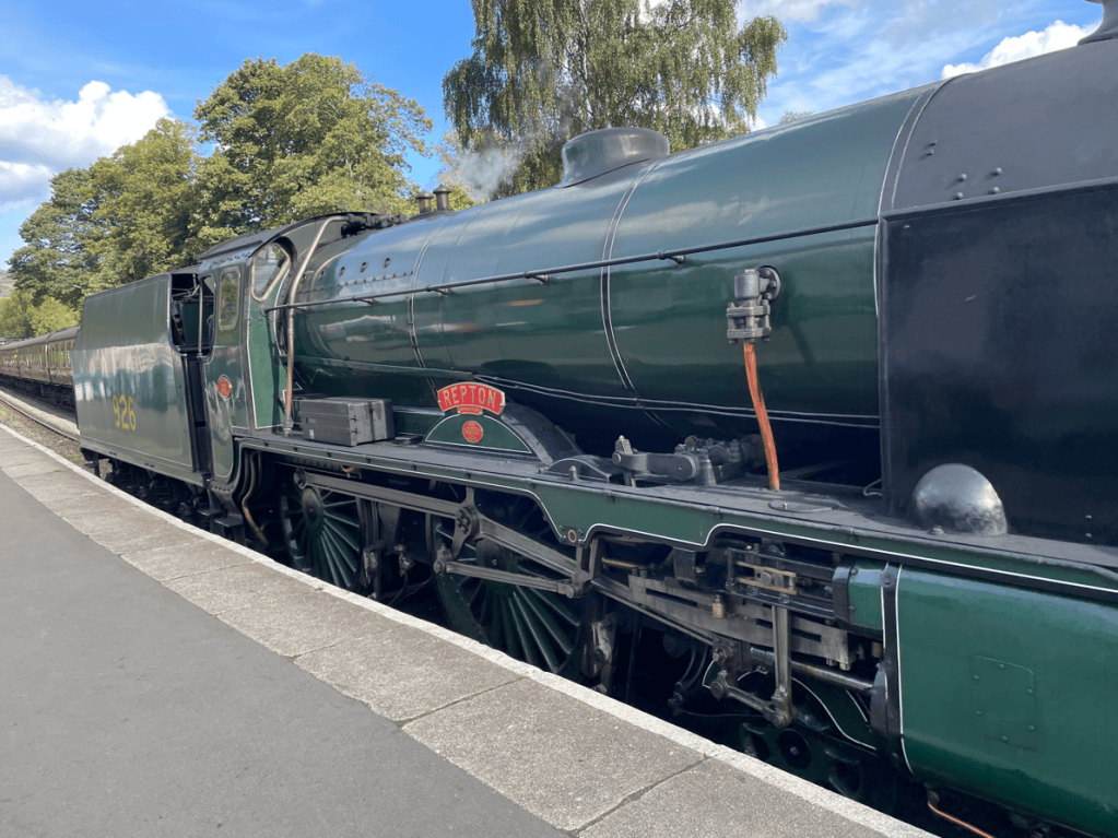 A green steam locomotive at a station with trees in the background and a blue sky.