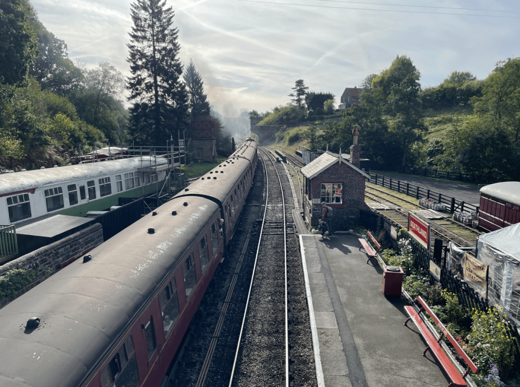 A view of a vintage train station with a red train in the foreground, surrounded by green trees and a hillside. The station features a small brick building and benches, with another train carriage in the background.