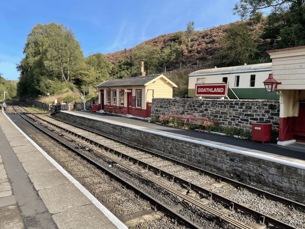Goathland railway station with wooden building and sign, surrounded by trees and hills, tracks visible in the foreground.