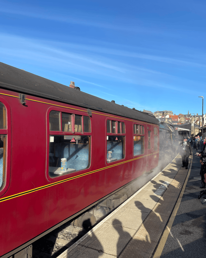 A red steam train at a station, with passengers on the platform and steam rising from the train.