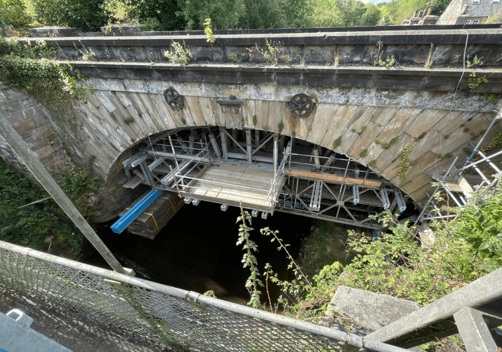 View of an arched stone bridge under construction, featuring scaffolding, wooden platforms, and overgrown vegetation on the sides.