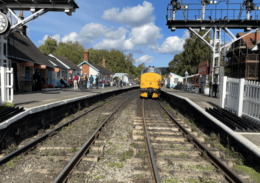 A yellow diesel locomotive at a railway station, with passengers waiting on the platform and historic station buildings in the background under a blue sky.
