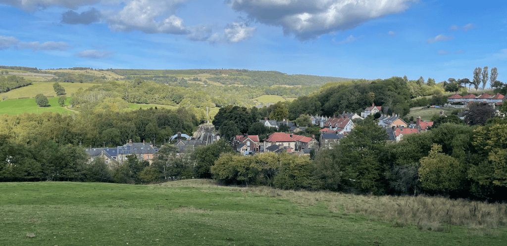 A scenic view of a small village nestled among green hills and trees under a partly cloudy sky.