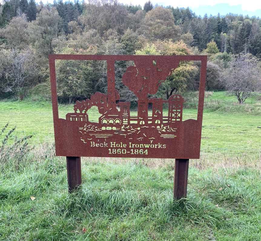A rusted metal sign depicting the Beck Hole Ironworks, highlighting its historical significance between 1860 and 1864, set against a backdrop of greenery and trees.
