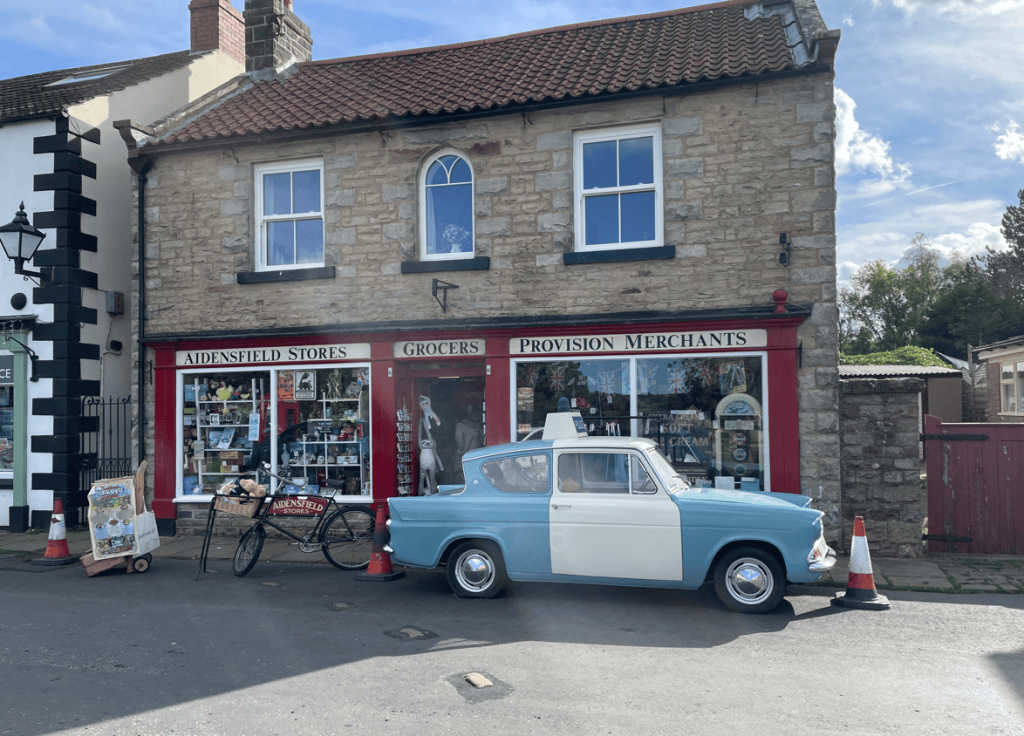 A quaint stone building featuring Aidenfield Stores and a grocer's shop, with a classic blue and white car parked outside and a bicycle nearby.