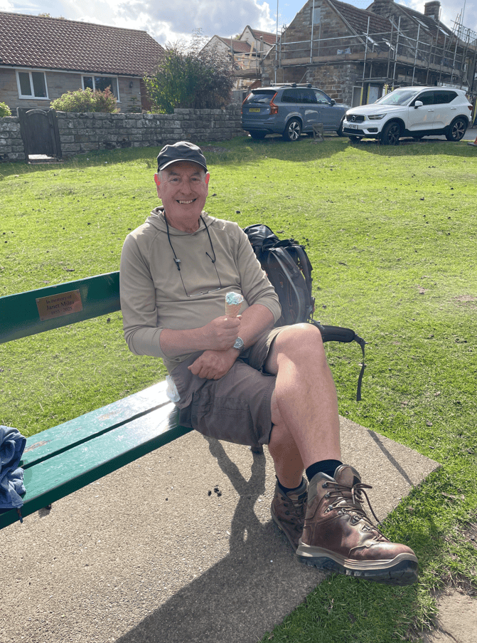 A man sitting on a green park bench, smiling and holding an ice cream, wearing shorts and hiking boots, with a backpack beside him, in a grassy area.
