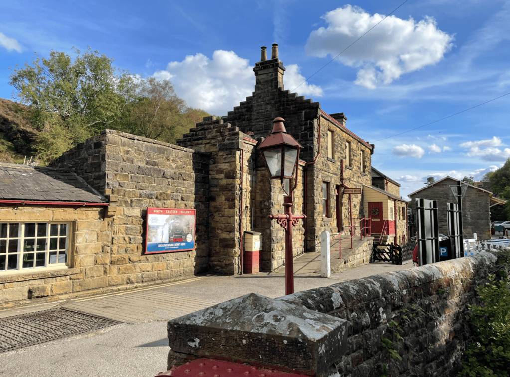 Historic stone railway station building with a red and grey façade, surrounded by trees and blue sky.