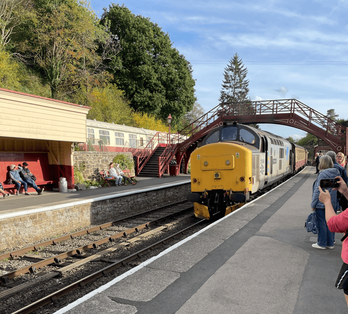 A yellow train at a station, with passengers waiting on the platform and an overhead bridge in the background.