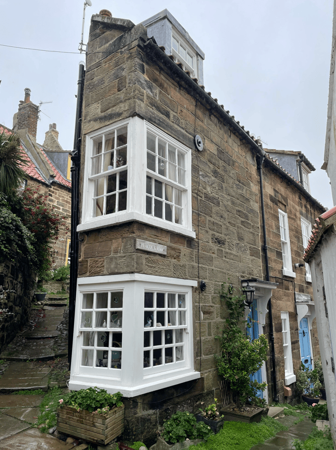 A quaint stone cottage with large white windows, located on a narrow, cobbled street. The façade features a decorative window bay, a small sign reading 'PEACEFUL ROW', and potted plants at the base.
