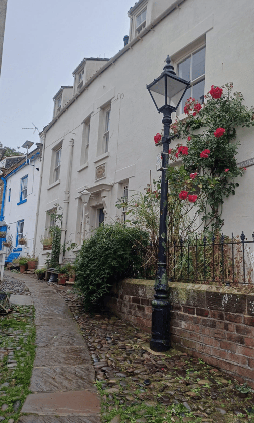 A narrow cobblestone path leading to a white building adorned with climbing roses and a vintage lamppost, with hints of blue and greenery in the background.