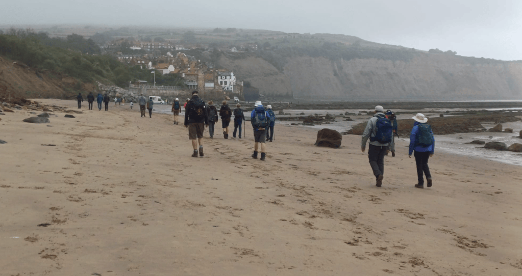 A group of walkers on a sandy beach, with coastal cliffs in the background and houses visible on the hillside under overcast skies.