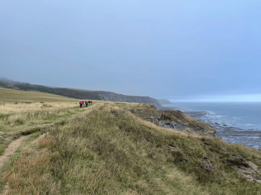A group of hikers walking along a coastal path with cliffs and the sea in the background, under a moody, overcast sky.