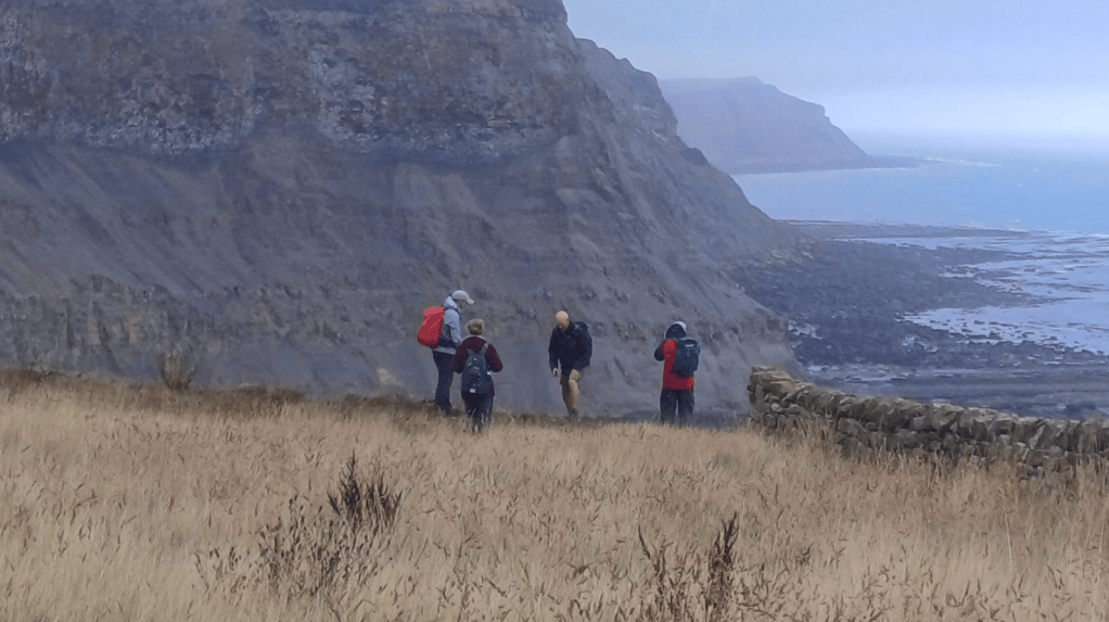 A group of hikers walking along a rugged landscape with cliffs and a coastline in the background. The terrain features dry grass and distant sea views.