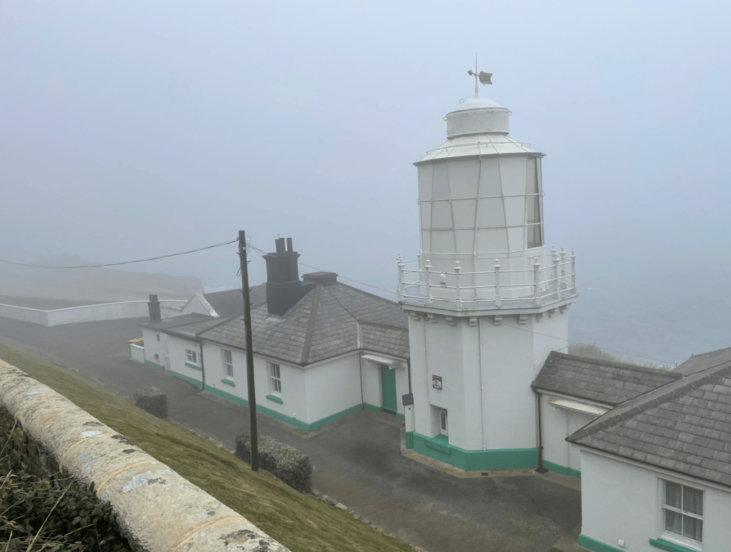 A white lighthouse surrounded by fog, with adjacent buildings featuring green accents and a coastal view in the background.