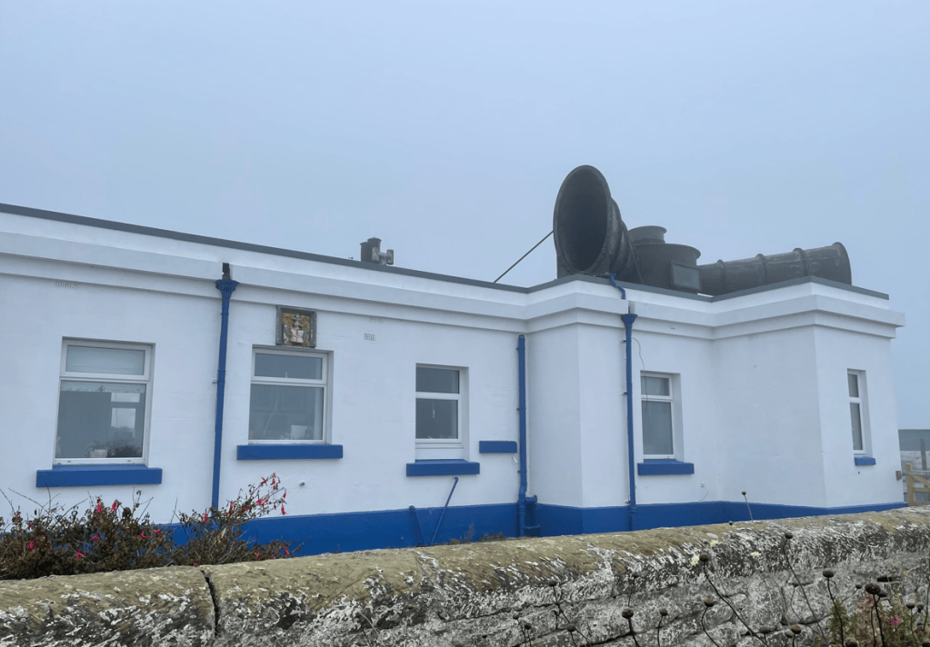 Exterior view of a white building with blue accents and a large black horn on the roof, surrounded by low fog.