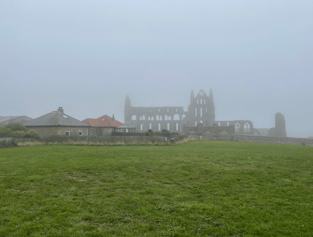 Ruins of an abbey partially obscured by fog, with nearby houses and green grass in the foreground.