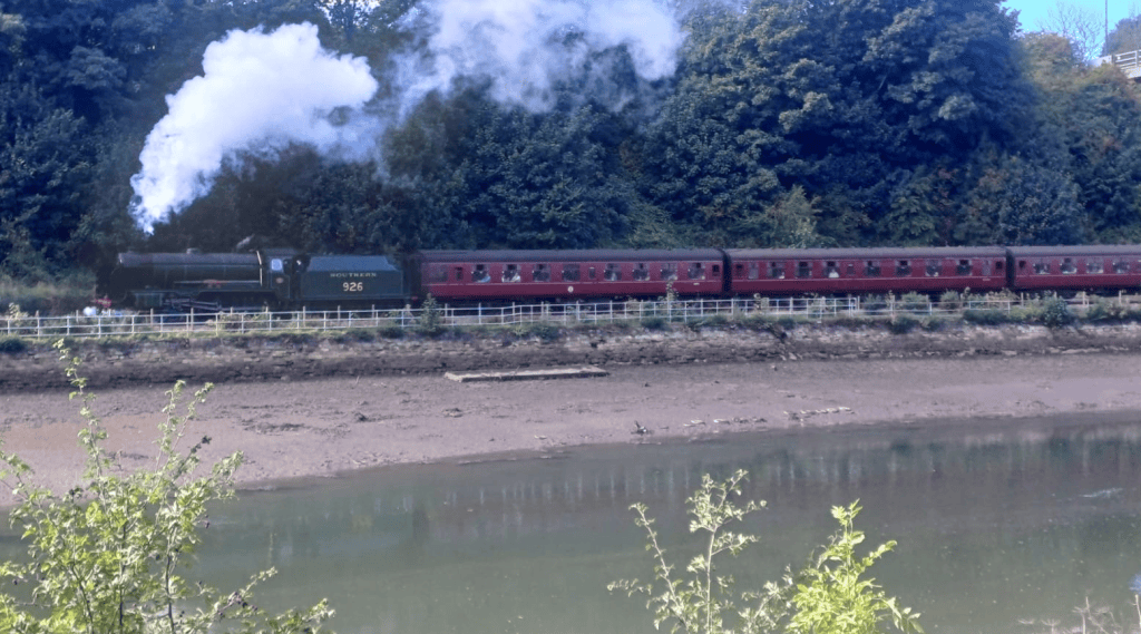 A steam locomotive numbered 926 pulling a series of red carriages alongside a river, with smoke billowing from the smokestack against a backdrop of green trees.