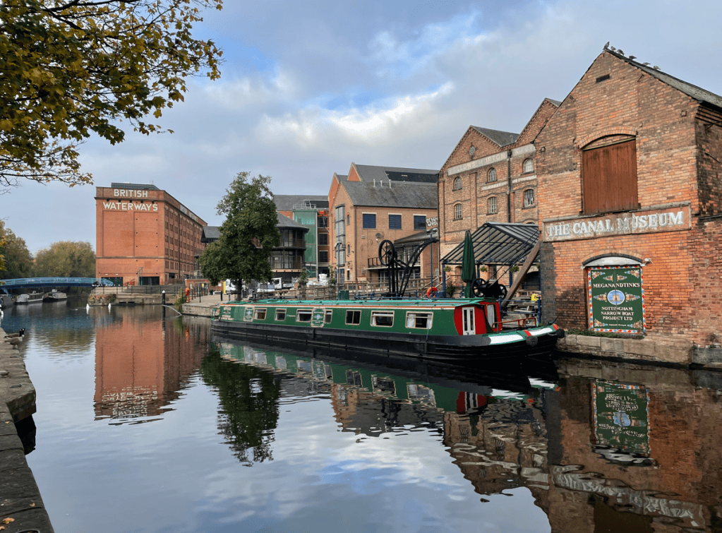 A narrowboat moored on a tranquil canal, reflecting the surrounding buildings, including a museum and historical brickwork, under a cloudy sky.