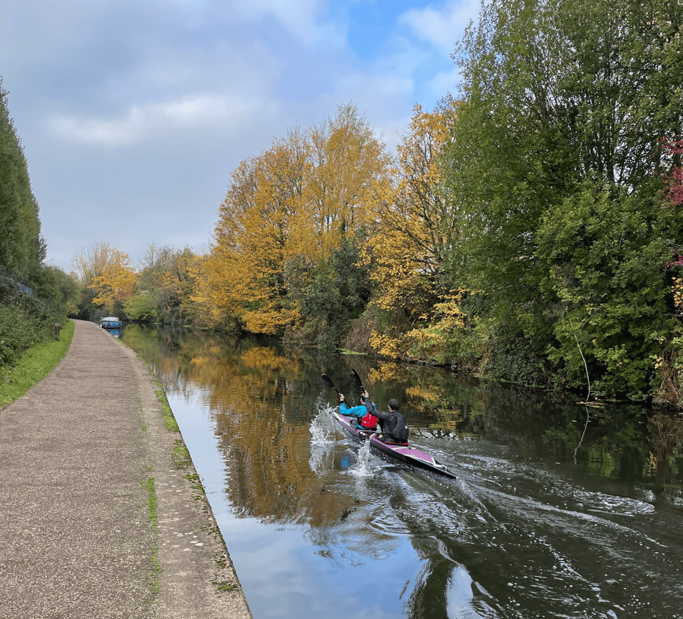 Two kayakers paddling on a serene canal with trees displaying autumn foliage along the banks.