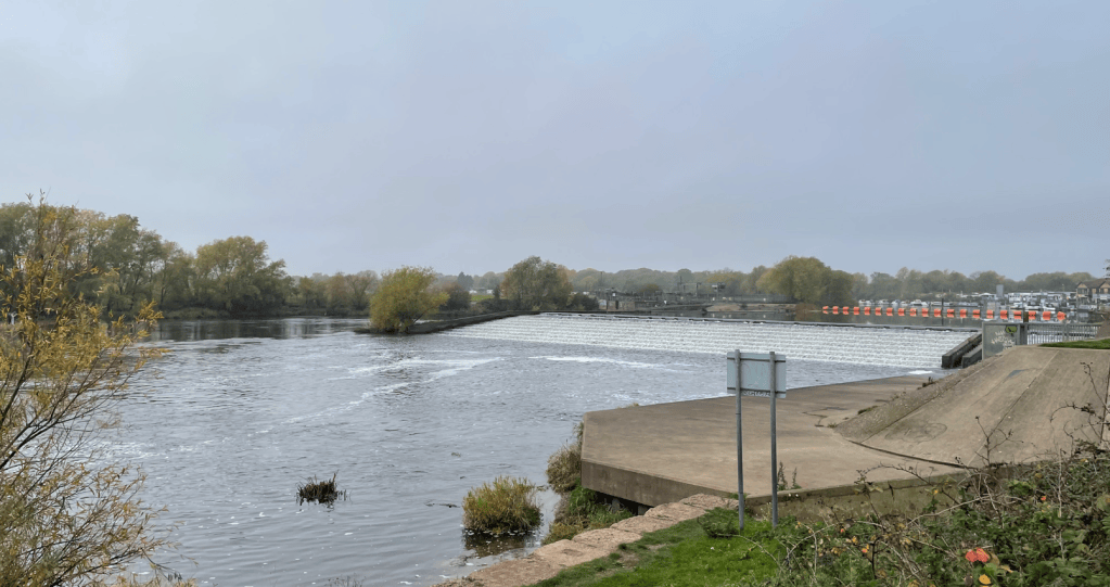 A view of a river with low water levels, lined by trees on either side, leading to a dam with a series of overflow cascades. In the distance, a row of orange barriers can be seen along the riverbank.