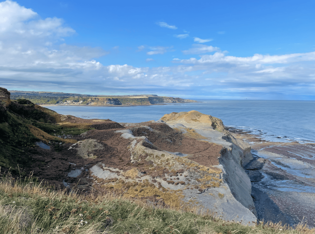 Coastal landscape showcasing cliffs, sand and rocky shore under a blue sky with scattered clouds.