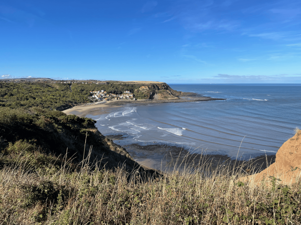 A scenic coastal view showing a sandy beach with gentle waves, surrounded by lush greenery and cliffs under a clear blue sky.