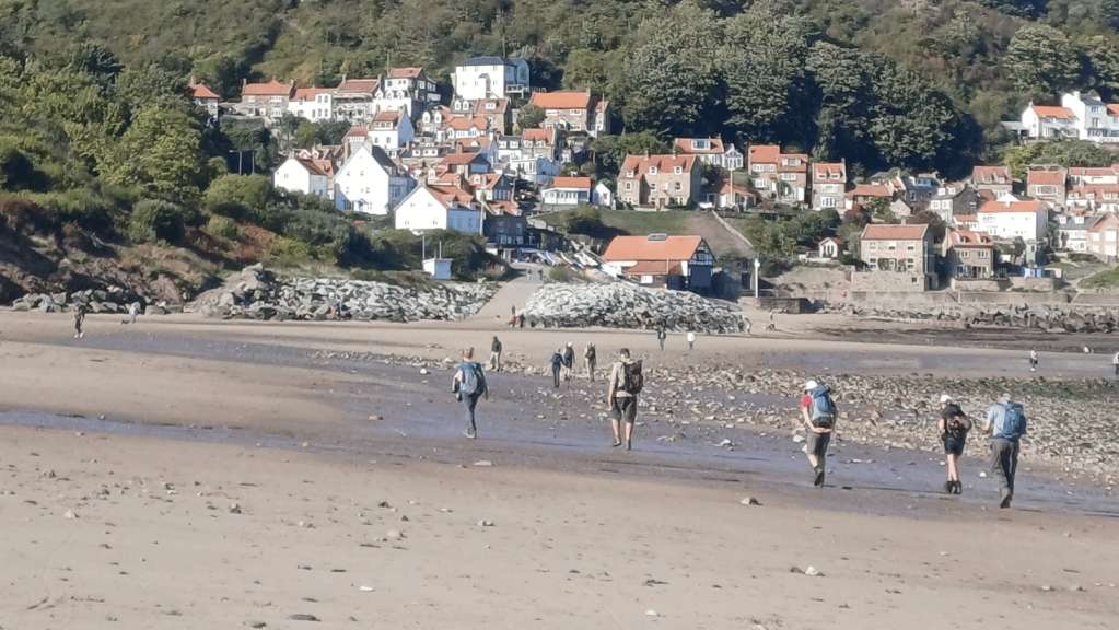 People walking on a sandy beach with a coastal village in the background, featuring houses with red roofs and greenery.