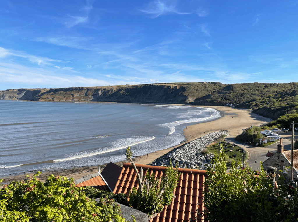 A scenic view of a beach with gentle waves and a rocky coastline, surrounded by lush green hills under a clear blue sky.