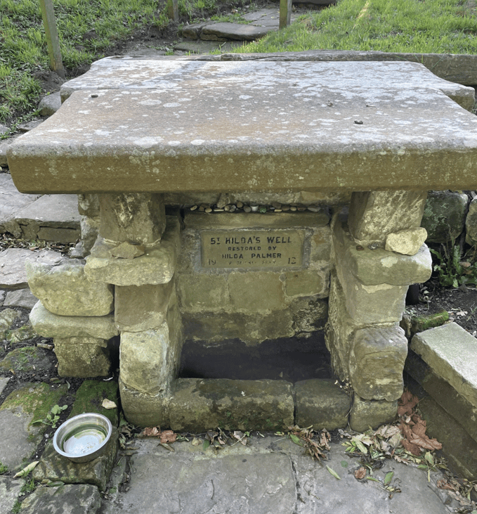A stone structure known as Hilda's Well, featuring a flat roof supported by stone pillars. A plaque with restoration details is visible on the front, along with a metal bowl and surrounding greenery.
