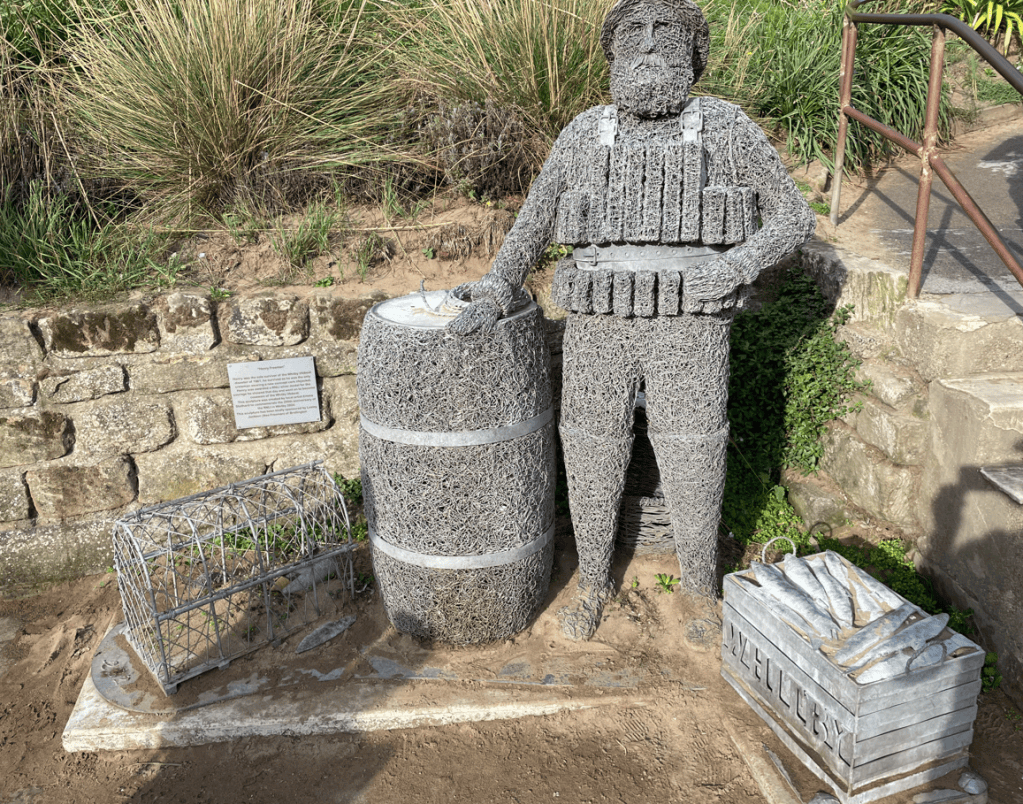 A wire sculpture of a man standing next to a barrel, with a basket of fish and a trap nearby, set against a grassy backdrop.